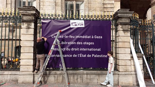Deux personnes renouvellent l'affichage mis en évidence sur les grilles de la mairie de Lyon (place de la Comédie). Sur cet affichage, le fond est violet et il y a, en haut, le logo de la Ville de Lyon. Puis le message en blanc "Cessez-le-feu immédiat à Gaza. Libération des otages. Respect du droit international. Reconnaissance de l'État de Palestine."