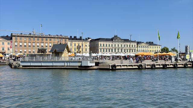A waterfront scene featuring a dock with a small building, busy festival tents, and a crowd of people. Historic buildings in the background display various architectural styles under a clear blue sky. Flags can be seen, adding to the vibrant atmosphere.