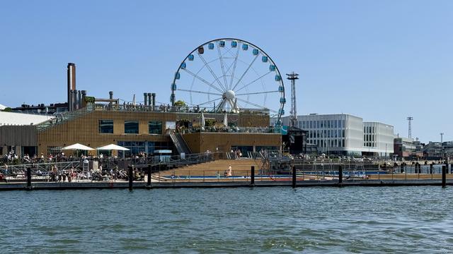 A lively waterfront scene featuring a large Ferris wheel, a modern wooden building with outdoor seating, and people enjoying the sunny day by the water. The background includes various structures, with clear blue skies above.