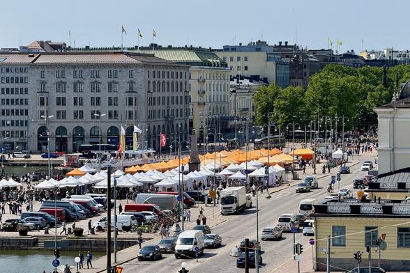 A busy street scene featuring multiple white and orange tents, likely for a market or festival. People are walking along the street and engaging with the stalls. Various vehicles are parked nearby, and a historic building with flags is visible in the background.