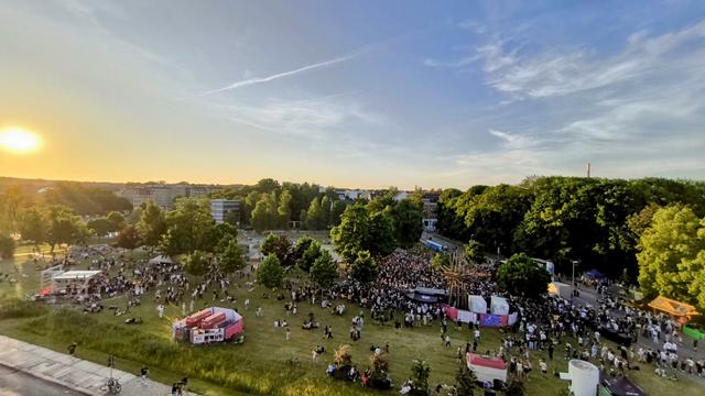 Blick von oben auf einen Park voller Bühnen, Stände und Menschen. Abendsonne scheint in die Kamera