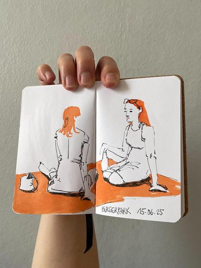 A hand is holding a small sketchbook with a 2 colored sketch. 2 girls in the park on a blanket.