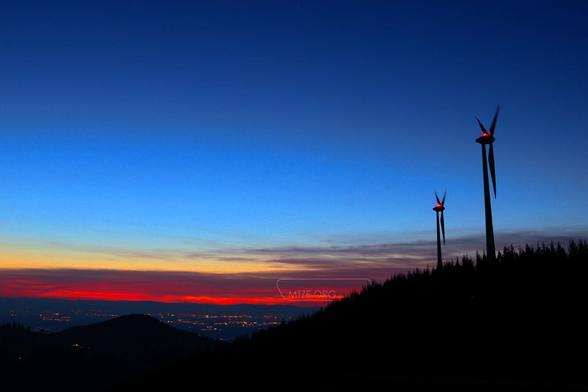 Eine Landschaftsaufnahme im Vordergrund sind ein schwarzer Berg mit zwei Windkraftwerken. Im Hintergrund ist ein Tal mit beleuchteten Gebäuden und einem Himmel in der blauen Stunde mit roten Wolken. / A landscape shot in the foreground is a black mountain with two wind turbines. In the background is a valley with illuminated buildings and a sky in the blue hour with red clouds.