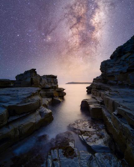 The vibrant core of the Milky Way galaxy rises from the horizon, framed between the dark, dramatic silhouettes of a rock quarry canyon. The stars glow in shades of purple and orange above a calm inlet of water that reflects the celestial light.