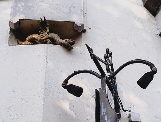 Witte muur met een uithangbord met lampje aan beide zijden. In een holte van de muur zit een stenen draak verborgen.