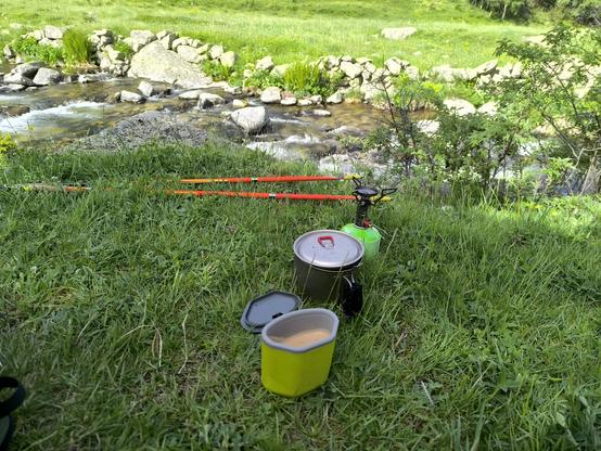 A green mug containing a hot drink stands on the grass in front of a titanium pot, which in turn is in front of a small stove with a green canister. The backdrop is a burbling stream.