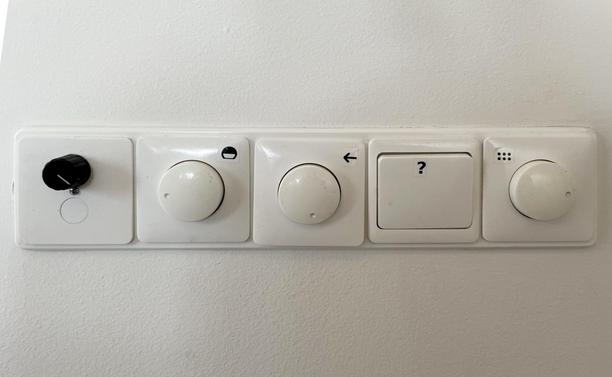 5 switches on a wall, left to right:

1. Small black with white button underneath
2. 3/4-sphere with black half and white second half
3. Arrow to the left
4. Question mark (yes)
5. 3x2 dots rectangle