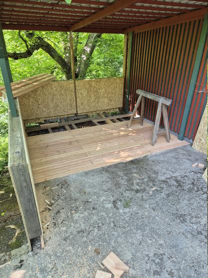 Eine Veranda ist im Werden. Ein Bretterboden, der zu zwei Dritteln verlegt ist und eine Brüstung, verkleidet mit OSB Platten, die von der linken Seite kommt und dann im rechten Winkel zur Gartenhütte auf der rechten Seite verläuft. Darüber befindet sich ein Dach aus Trapezblech.