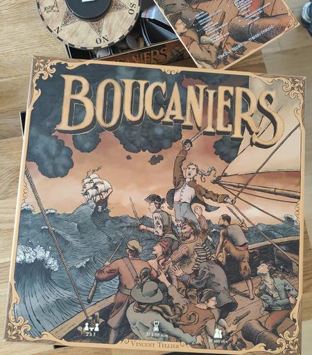 Boite du jeu Boucaniers, avec un groupe de pirates se galvanisant à aborder un autre navire au loin. Les vagues sont furieuses, le ciel orange et quelques nuages sont noirs et menaçants.