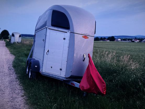 A silver horse trailer is parked on a gravel road next to green fields. A red flag is attached to the rear of the trailer. The sky is slightly overcast, suggesting twilight or dusk. In the background, a second horse trailer is visible along