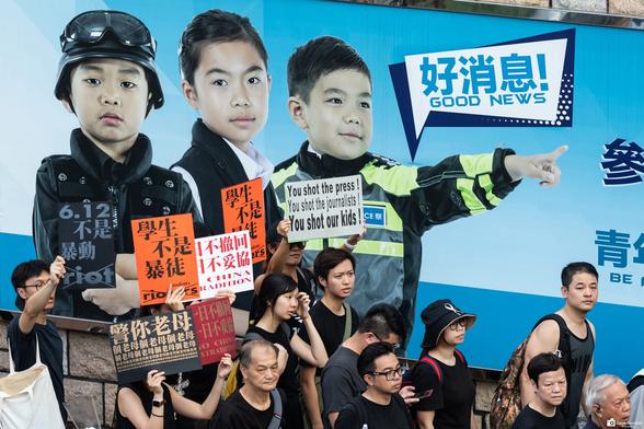 Protesters marching past HK Police headquarters, with a large police recruitment advertisement on the wall while carrying placards complaining about police brutality.