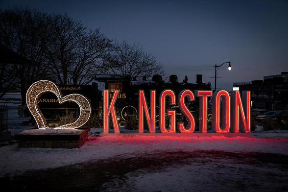 The illuminated Kingston, Ontario sign glows warmly at night, enhancing the winter scene with a charming heart symbol and historic train backdrop.