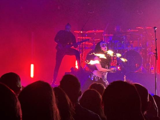 Ne Obliviscaris bass player Martino Garattoni playing on a stage saturated in red light, while fire artist Jessie Spins twirls a frame with five points of flame past her face.