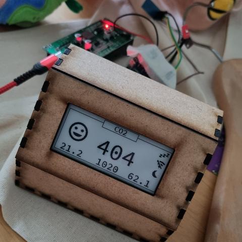 Ein Holzgehäuse, in dem ein epaper Display steckt. Auf dem Display steht oben CO2, groß in der Mitte die Zahl 404, auf der linken Seite ein fröhliches Emoji. Darunter sind mehrere Zahlen, am rechten Rand einige Symbole. 
Im Hintergrund sind elektronische Bauteile zu erkennen.