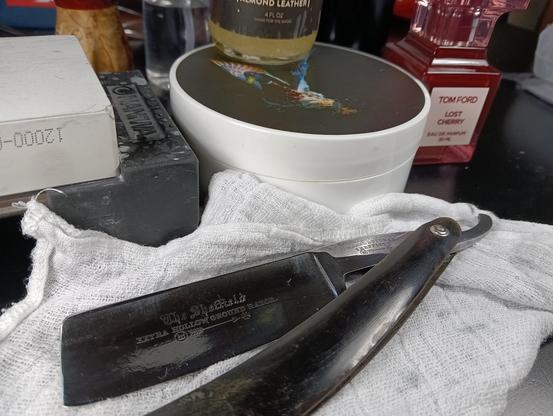 (Le Chat Mistral description)

The image depicts a traditional barber setup. At the centre, there is a white ceramic bowl with a liquid, possibly for shaving. A bottle labelled "Balmond Leather" is placed above the bowl. To the right, there is a straight razor with a dark handle, resting on a white cloth. The razor has "The Sheffield Extra Hollow Ground Razor" inscribed on the blade. Nearby, there is a black whetstone for sharpening the razor. In the background, there is a red bottle of Tom Ford's "Lost Cherry" perfume. The scene is set on a dark surface, giving it a classic and professional feel.