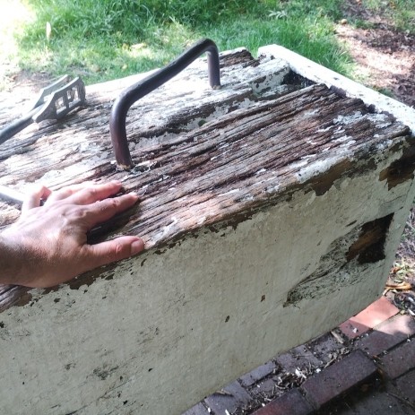 A man's hand and a windlass laid atop the end of a square-cut oak beam beside a steel grab handle.

This photo focuses on the end of the beam. The beam is part of a massive oak lock gate (not shown).

A lock beam is a lever that lets boaters open a wooden lock gate that can range from a very small 750 kilos to a typical large size of 1500 kilos (about ¾ to 1½ tons). This beam is probably 50×50 centimetres or 20×20 inches—and has a flat end. Its full length isn't shown in the photo but might be 7 meters or 22 feet. The very end of the beam is bound by a reinforcing, flat steel bar, to keep it from splintering.

There's a solid steel grab handle anchored in the top surface of the beam, for boaters to grab and pull, to start the gate moving. The end of the beam is painted white, though the paint has worn away on top, as has the wood, from years of use and from getting tapped by windlasses. (Boaters usually have those in one hand as they reach for the grab handle with both hands.)

The windlass in the photo is a type of spanner or wrench, one piece of steel, with a square hole on one end that fits the lock mechanism. (Mechanism not shown.) For easier winding, each side of the windlass' L-shaped handle is about 30 centimeters or 1 foot, and about 2 centimeters or ¾ inch round in diameter.

In the background is a dirt towpath and some unmowed grass. Below the beam is red-brown brick paving with some ridges set in to offer some traction to boaters as they strain to push the beam.