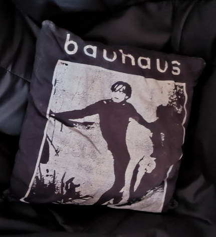 Un cojín de tela oscura sobre un puff de polipiel negro. El cojín tiene una imagen en blanco y negro de una persona con una postura estirada, con otra figura menos definida a su lado. El nombre "Bauhaus" está impreso en la parte superior del cojín en letras blancas grandes. Laimagen pertenece a un fotograma de la película muda "El Gabinete del Doctor Caligari"