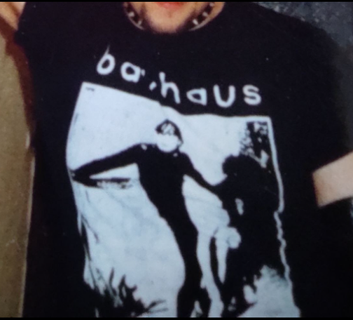 Una camiseta negra muestra un diseño en blanco y negro de la banda Bauhaus. El nombre de la banda está impreso en la parte superior del diseño. Debajo, una imagen en blanco y negro, estilo silueta, representa dos figuras humanas en un abrazo o interacción física cercana. El estilo visual es sencillo y un poco oscuro, acorde con la estética gótica de la banda.