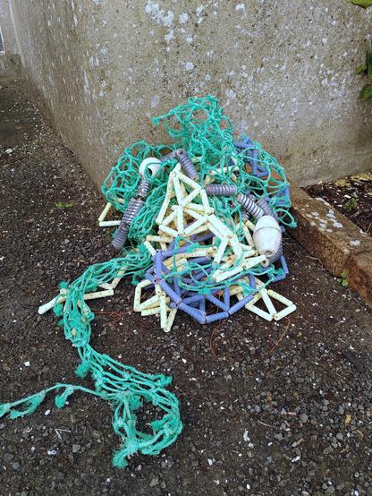 Ceramic floaters, lines and ceramic fishing net combine with real fishing net parts laying on the concrete in a messy stack against a cement wall.