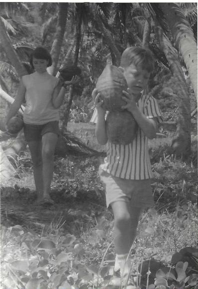 A black and white photo shows a boy and young woman carrying large coconuts in a tropical setting with tall palm trees and dense foliage.
