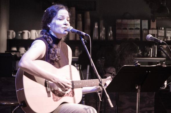 A woman playing guitar and singing into a microphone at a coffeehouse. She is mostly on the left side of the frame; in the background are coffee mugs and boxes of tea on shelves.