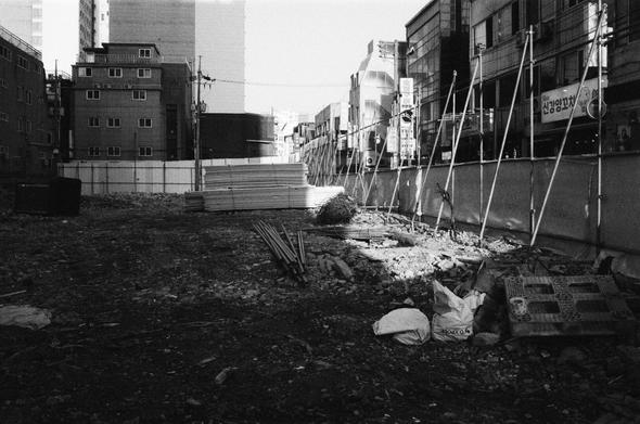 도심 속 폐허. 펜스로 둘러싸인 채 건물 잔해, 공사 자재, 쓰레기가 널려있다. 주변 건물들이 둘러싼 것처럼 보인다.