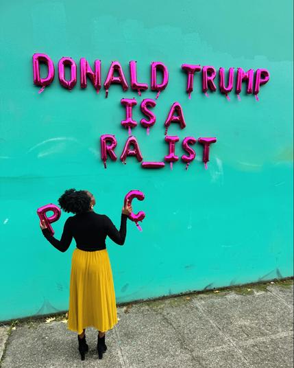 « Trump is a ra_ist » etcrit sur un mur bleu pâle. Devant une femme noire, de dos, tient à la main les lettres « P » et « C ».