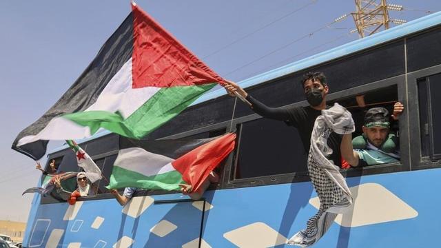 Fotografía de un autobús azul con detalles blancos y ventanas negras. De las ventanas del camión se asoman activistas de la Marcha Global a Gaza ondeando banderas de Palestina.