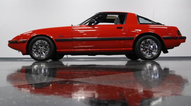 Photo of an early 1980s FB Mazda RX-7 in red taken from direct left side low. Car is in a white room with a black reflective floor and the overhead lights are reflecting nicely on the car's paint