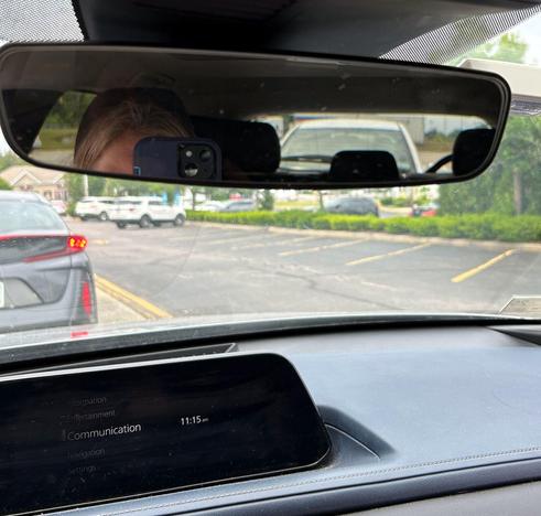 a person with blonde hair and light skin tone taking a photo in the rear view mirror of their car. A white car is visible in the mirror, a black car through the front window.