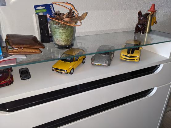 Die Oberseite eines Schuhschranks. Unter der Glasplatte stehen drei Fahrzeugmodelle: Golf 1, Porsche 959, Chevrolet Camaro (Bumblebee)