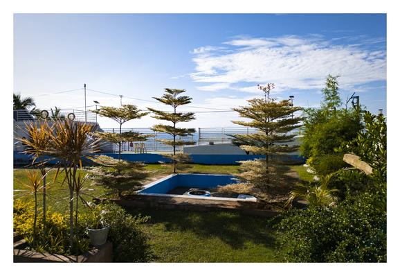 A sunlit garden with green grass, various plants, and a small fire pit is pictured in the foreground. In the background, a structure resembling a boat deck with white railings overlooks the ocean under a bright blue sky with scattered clouds. - Google Gemini Pro 2.5