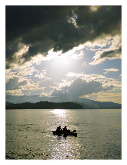 A dramatic, backlit shot shows two people in a small boat on a shimmering body of water. Above them, the sun breaks through dark, heavy clouds, with hazy mountains visible in the distance.
