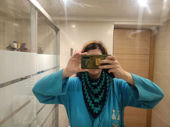 Selfie de espejo de baño, pero anonimizado por ponerme el móvil a la altura de la cara.
Lo importante es el pañuelo que llevo en torno al cuello y que cae sobre el pecho alternando franjas verde azuladas y negras.
Llevo puesta la cinta del pelo moebius naranja que me hice al principio del cursillo de ganchillo.
Visto una bata ligera azul de la que no me preguntéis por qué tiene el bordado de un bikini, porque no conozco la respuesta e_e