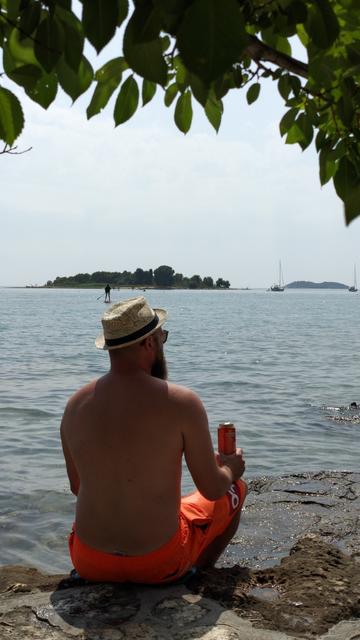 Mann mit Bart, Hut und einer Dose Bier in der Hand ist von hinten zu sehen, er sitzt auf einem Stein am Ufer des Meeres, darüber ein paar Äste und Blätter eines Baumes, im Hintergrund eine kleine Insel