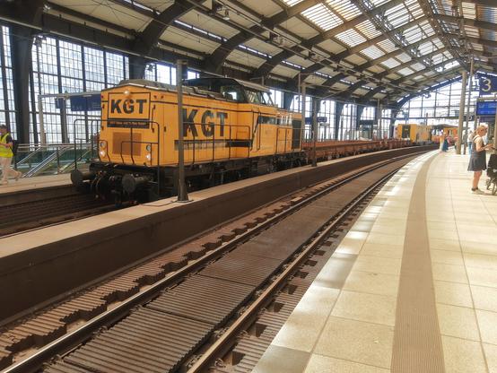 Das Bild zeigt die Bahnhofshalle Friedrichszraße mit einer gelben Bauzug-Lokomotive. Die Lokomotive trägt die Abkürzung "KGT" auf ihrer Seite. Die Halle hat ein hohes, offenes Dach mit großen Fenstern, die viel natürliches Licht hereinlassen. Auf der rechten Seite der Gleise befindet sich eine Plattform, auf der einige Personen stehen und warten. Einige der Personen tragen Sicherheitswesten. Im Hintergrund sind weitere Gleise zu sehen.

Bereitgestellt von @altbot, privat und lokal generiert mit Ovis2-8B