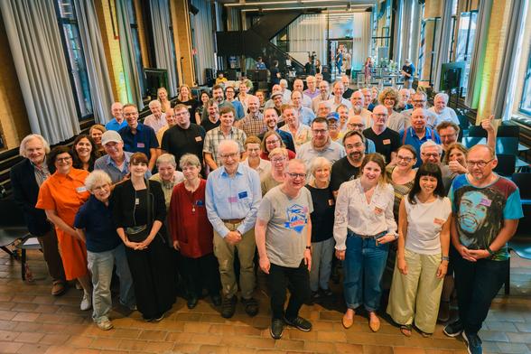 Zu sehen ist eine große Gruppe an Menschen, in etwa 50-60 Personen. Diese stehen in der Mitte eines großen Raums und lachen in die Kamera.