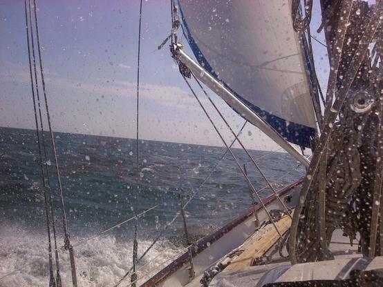 Color image from the aspect of the cockpit of a blue-water sailboat plying the Atlantic Ocean off Coastal Carolina, sails full, ocean blue and inviting, clouds on the horizon.
