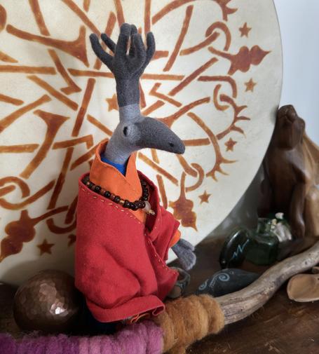 a needle-felted monk creature on a wooden bookshelf. the monk has a dark grey curved beak, and some dark grey antler-type things on top of their head. they have kind, dark eyes on their lighter grey head. they're wearing robes in red and orange with a necklace of amber-coloured beads around their neck, and you just just see some bright blue poling out from their collar. one of their hands is outstretched slightly. they have an aura of calm wisdom and kindness. 
they're standing in front of a large drum decorated with henna patterns. there's a hammered copper ball next to them and an oak stick wrapped in plant-dyed wool in front of them: the colours you can see here range from dark to light pink and dark to light orange. there's a wooden hare sculpture leaning slightly against the drum on one side. it has a small lily-of-the-valley flower fairy at its feet along with a green glass bottle. next to that is a medicine bear carving, and next to that is a stone in the shape of a whale or dolphin painted with dot and swirl designs. there's also a tiny ceramic bowl and the tip of a welsh love spoon. and i'm glad i cropped the photo here or i'd be typing all day!