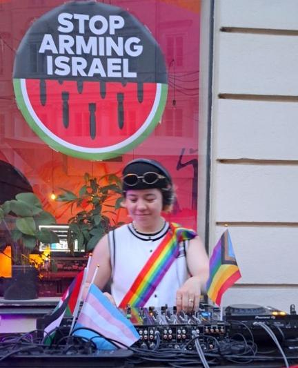 A DJ with equipment with a LgBTQ+ flag, behind a banner "stop arming Israel" with a water melon