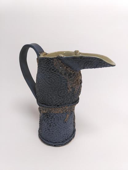 Handbuilt matte black ceramic pitcher with hammered like textured that are usually found on metallic object. some part look kind of rusty from a metallic glaze used on it.