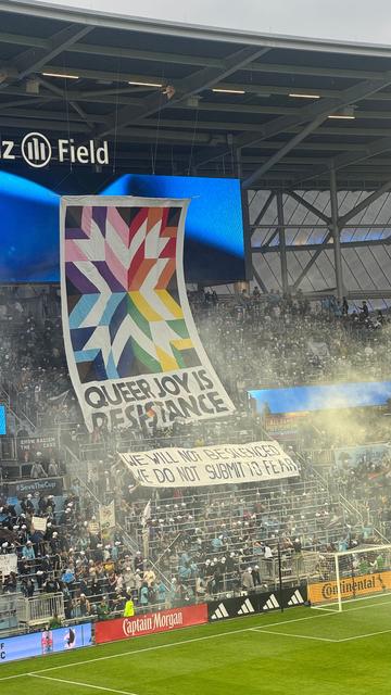 Huge banner hoisted up by the crowd at a soccer game, with the words “QUEER JOY IS RESISTANCE” and a smaller banner that reads “we will not be silenced, we do not submit to fear”