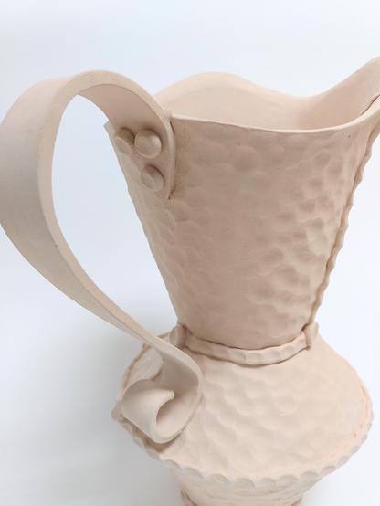 Bisque fired handbuilt pitcher. Handbuilt. It has a hammered like texture like we can find on hammered copper objects. it is built out of 3 conical parts. 2 cones are creating the body and the third creates a large spout opening. big flat handle.