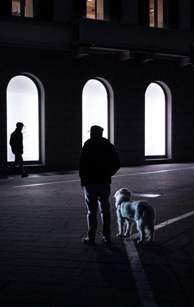 Night. A man with a dog stands and seems to wait on the dark street. His silhouette stands out in front of a bright shop window. Another man walks past a similar shop window.
