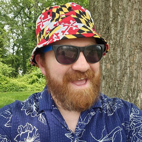 Selfie of a bearded, light skinned man in a blue shirt with a white floral pattern. The man is wearing sunglasses and a bucket hat covered in a Maryland state flag pattern.