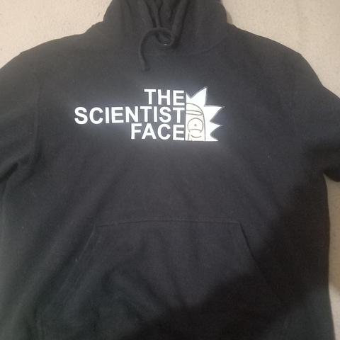 La imagen muestra un polerón (sudadera con capucha) de color negro. En el pecho, tiene un diseño en blanco que es una parodia del logo de "The North Face". En lugar de "The North Face", se lee "THE SCIENTIST FACE", y el logo con el arco que normalmente representa una montaña, está modificado  la cabeza de Rick Sánchez, el personaje científico de la serie animada "Rick y Morty", con su icónico cabello puntiagudo.
