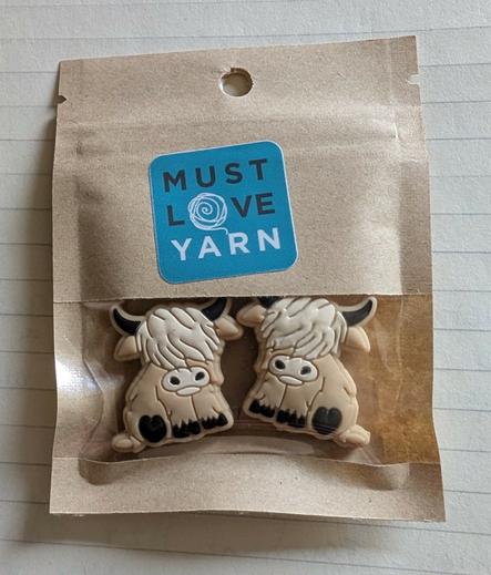 Two knitting needle point protectors in the shape of tan Highland cows. In a package that has the words "Must Love Yarn" on it.