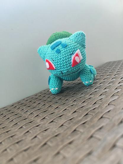 A handmade crochet Bulbasaur, crafted using teal and green yarn, sits against a neutral background. The details of the amigurumi, including its wide eyes and leafy bulb, showcase intricate needlework.