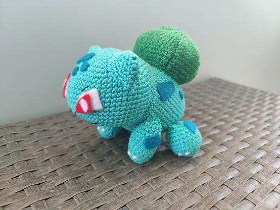 A handmade crochet Bulbasaur plush toy rests on a woven surface. The light teal body features dark teal spots, a leafy green bulb on its back, and striking red eyes with white accents. The detailed needlework highlights the craftsmanship in replicating this Pokémon character.