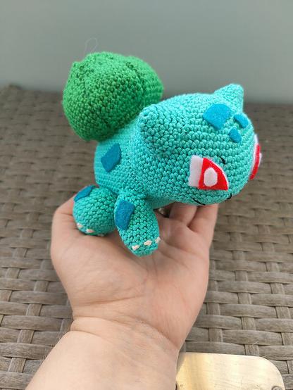 A hand gently holds a crocheted Bulbasaur plush toy, showcasing its detailed craftsmanship. The toy is made from light teal and green yarn, with red eyes and a leafy bulb on its back. The woven background adds texture to the scene.